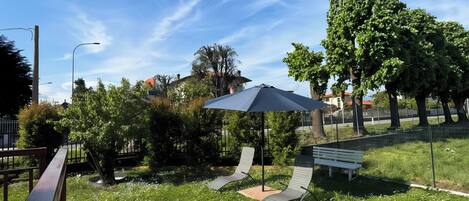 A grassy outdoor area with a stone pathway, a white bench, and two lounge chairs under a blue umbrella.