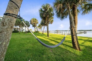 A hammock tied between trees in a grassy area with a body of water and a dock in the background. - Laguna Bella C4 (Gulf Shores)