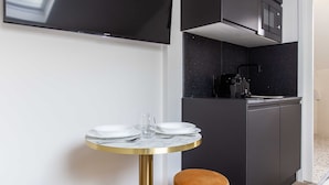 A modern hotel room with a round dining table, two ottomans, a flat-screen TV, a kitchen area, and a bed. - Cosy Studio - 2P - Parc des Expositions (Paris)