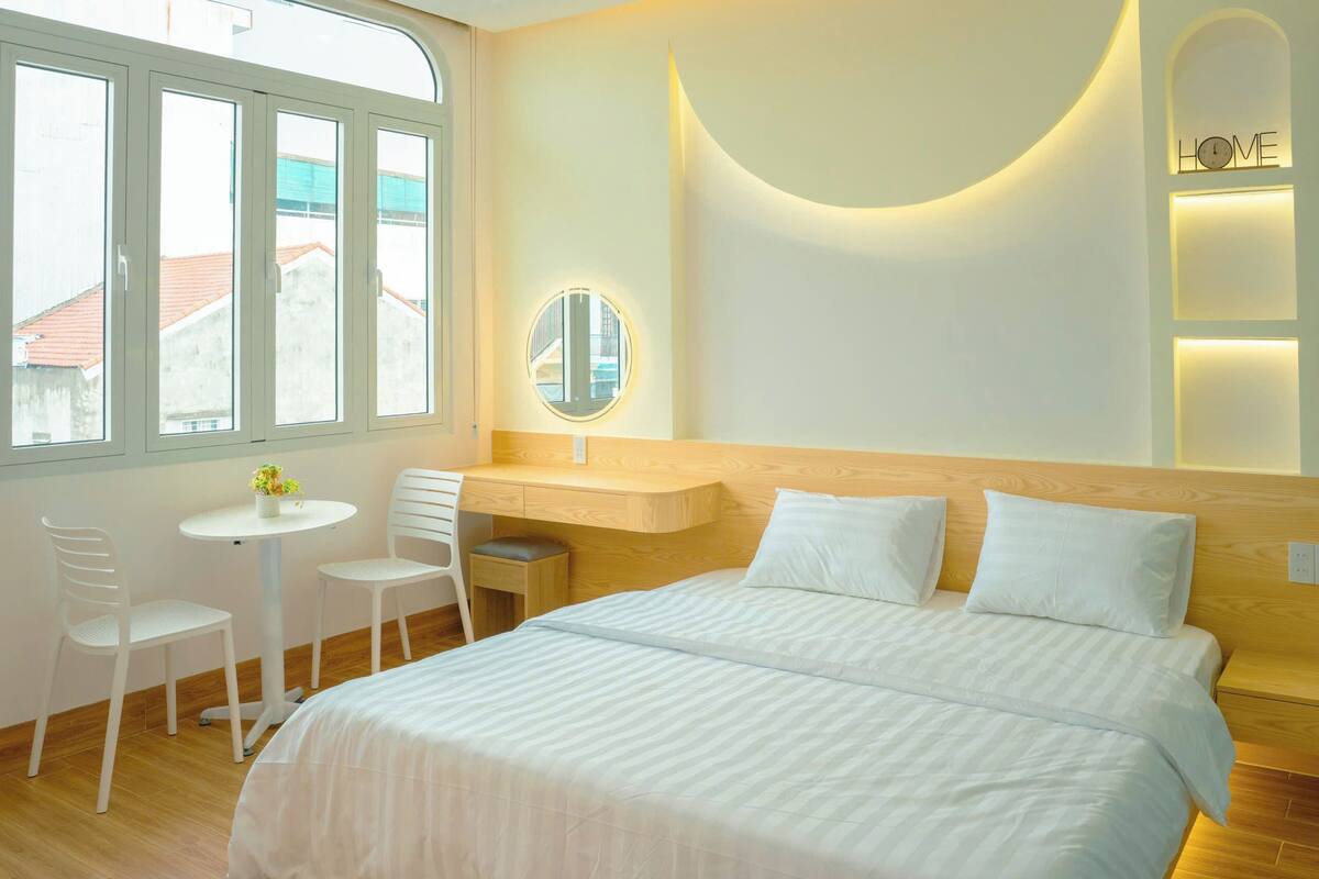 A hotel room with a bed, a desk, two chairs, a small table with flowers, a round mirror, and a view of buildings outside the window.