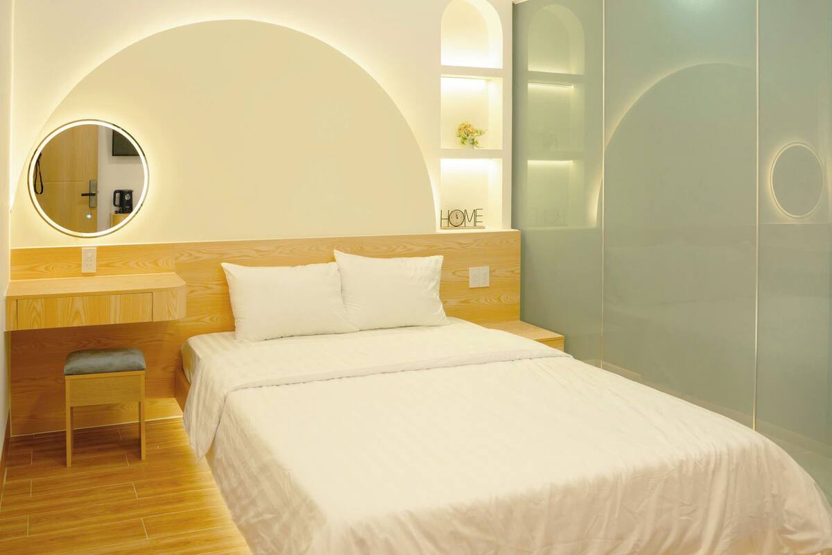A bedroom with a bed, a small stool, a round mirror, and a wall-mounted shelf with a vase.