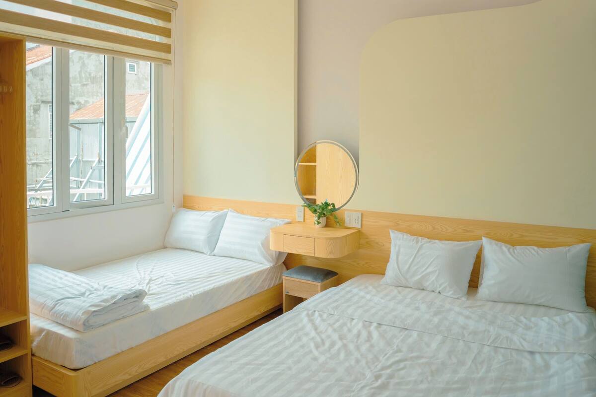 A hotel room with two single beds, a small bedside table with a mirror, and a window with a view of buildings.
