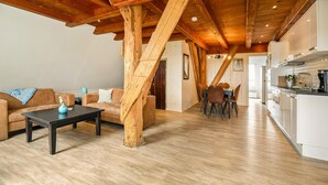 Un vaste espace de vie doté d’un plafond en bois, d’une table à manger avec des chaises, d’un canapé et d’un coin cuisine avec des meubles blancs.