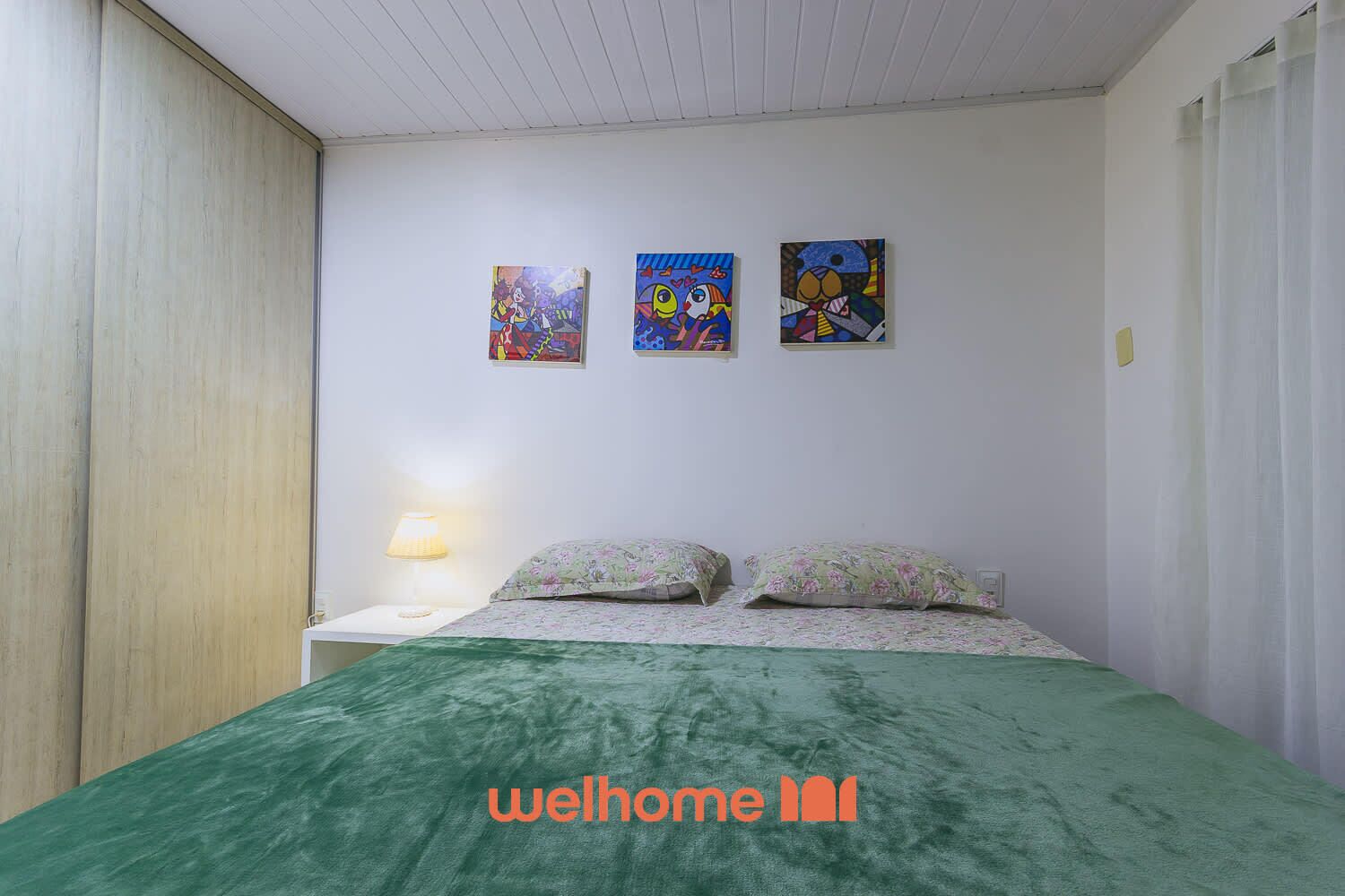Ein Schlafzimmer mit grüner Bettdecke, zwei floralen Kissen, einem Nachttisch mit Lampe und drei farbenfrohen Bildern an der Wand.