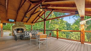 A wooden deck with a stone fireplace, outdoor seating, and a view of the forest. - Hesed Retreat | Views + Secluded + Hot Tub (Gatlinburg)