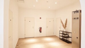 A spacious room with wooden flooring, white walls, and three white doors. A fire extinguisher is mounted on the wall, and a black shelf with pampas grass is placed against the wall. - Hotel Kronprinz  (Emden)