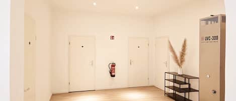 Une pièce spacieuse avec un plancher en bois, des portes blanches, un extincteur et une console noire ornée de quelques branches de graminées des pampas.