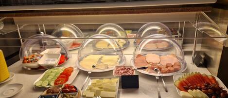 Buffet com diversos alimentos sob capas de vidro, incluindo queijos, carnes e frutas.