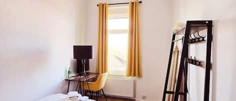 Una habitación pequeña y bien iluminada con una cama, un escritorio con silla, un radiador y una ventana con cortinas.