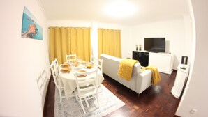Un coin repas avec une table blanche dressée pour quatre personnes, des rideaux jaunes et un canapé blanc agrémenté d’un plaid jaune.