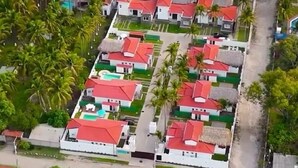 An aerial view of a coastal resort with rows of houses with red roofs, surrounded by palm trees and beachfront access.
