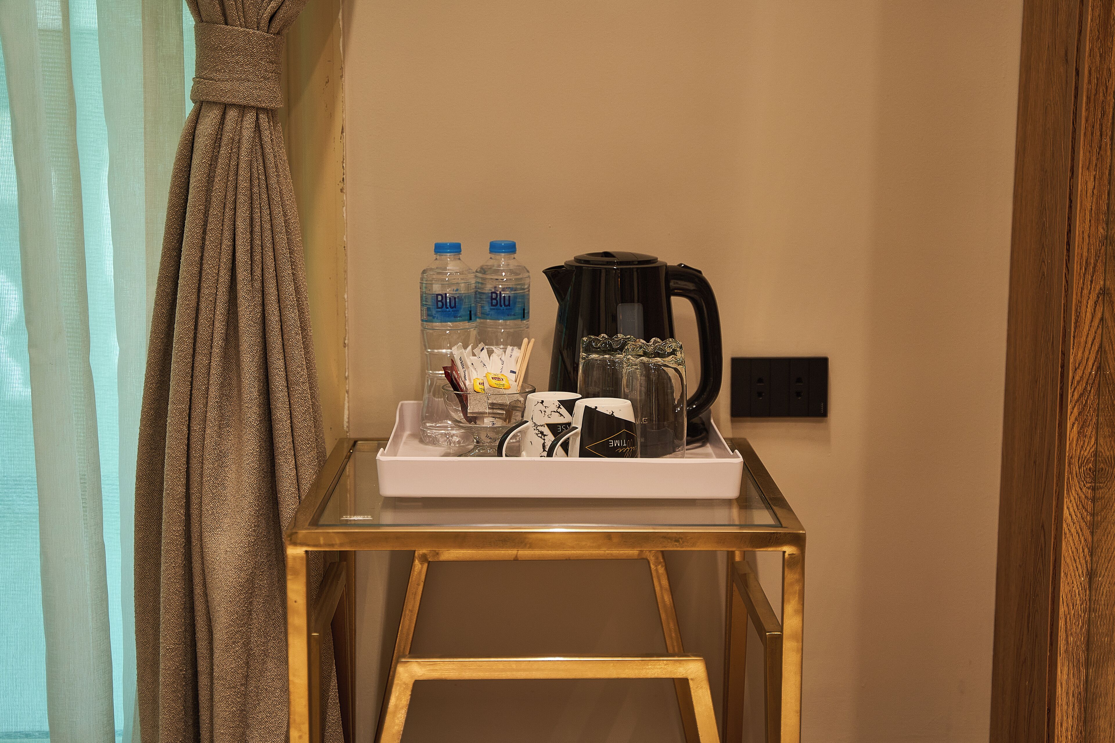 A hotel room with a glass table holding a tray with a coffee maker, two water bottles, a mug, and a small bowl.