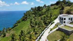 A modern building with a white exterior, surrounded by lush greenery and palm trees, overlooking a coastline. - Mountaintop villa for film, TV and photo shoots only.  Not for vacation rental. (Samana)
