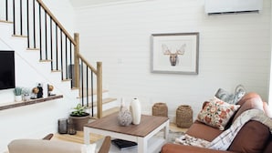 Un salon moderne avec un escalier en bois, un téléviseur fixé au mur, une œuvre d’art représentant une tête de cerf encadrée et un canapé marron confortable agrémenté de coussins à motifs floraux.