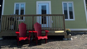 Une maison verte avec un toit triangulaire, une véranda avec des fauteuils Adirondack rouges et un ciel bleu azur.