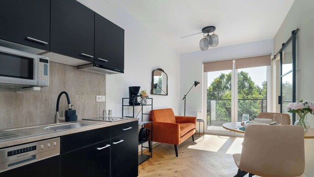 A modern kitchen with black cabinets, a built-in microwave, and a dishwasher. There is a dining area with a round table, orange sofa, and a chair. A large window provides a view of the outdoors.