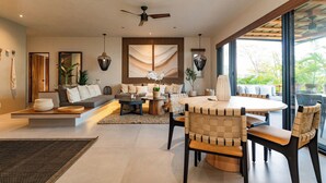 A modern living room with a sofa, coffee table, and dining table. There is a ceiling fan, large windows, and a view of greenery outside. - Stylish Escape | Private Plunge Pool | Oceanviews (Huatulco)