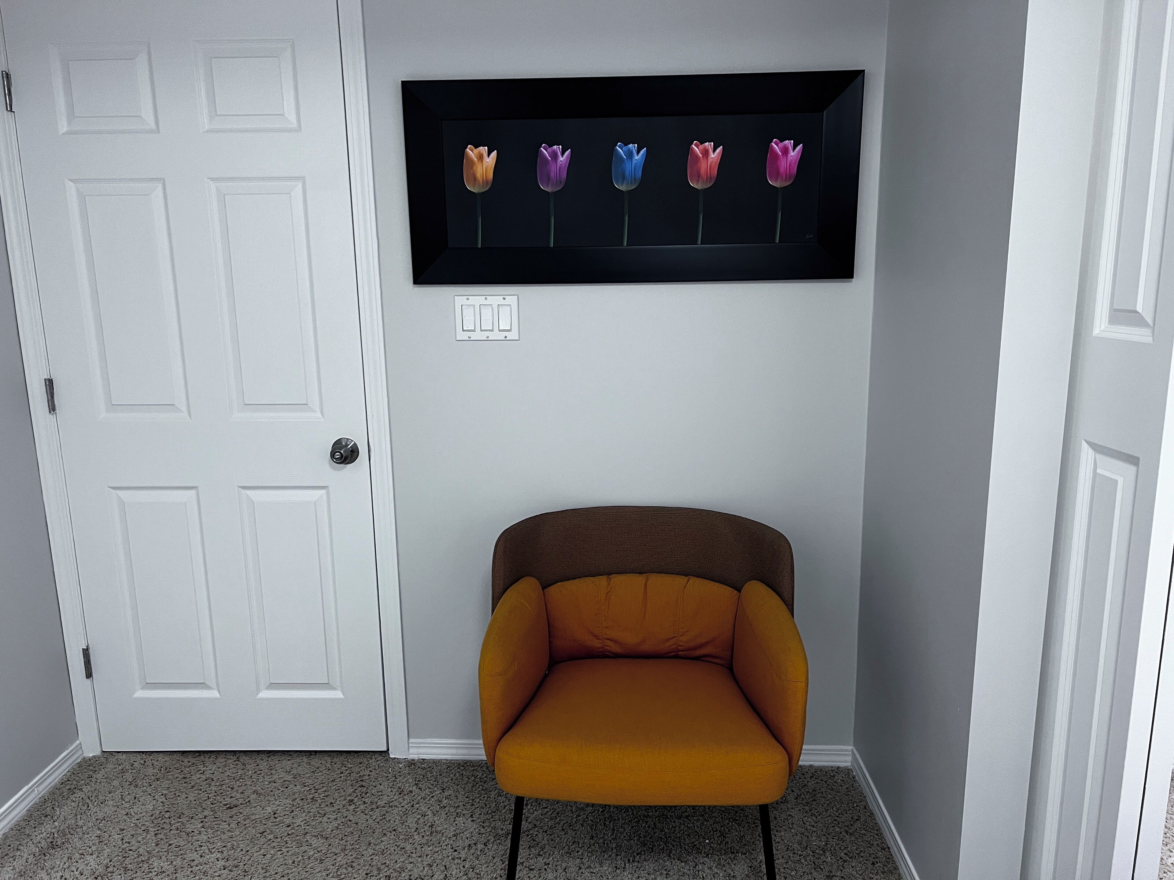 A room with a white door, a framed picture of flowers on the wall, and a mustard-colored chair.