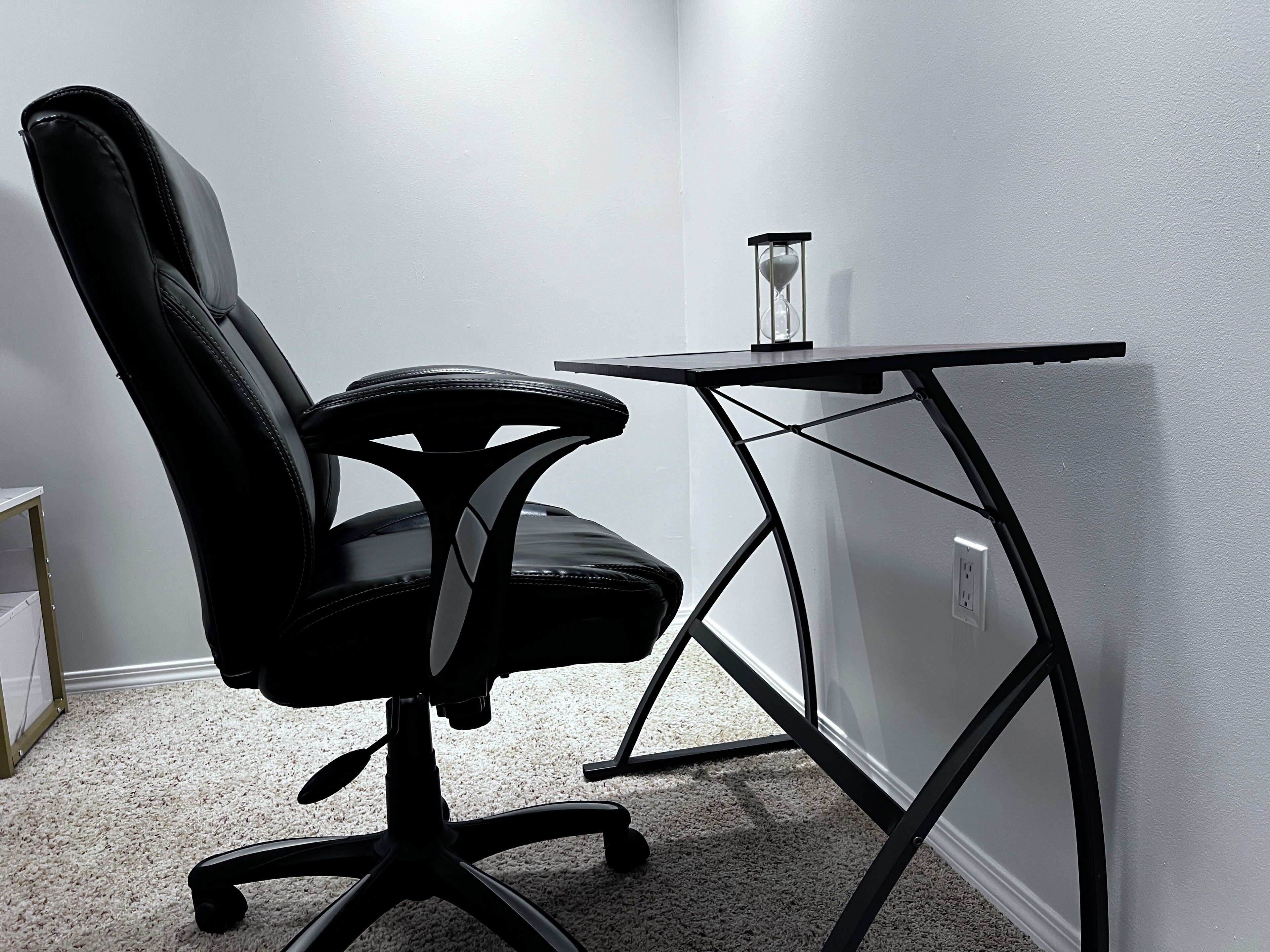 Un bureau d’angle avec un fauteuil de bureau noir, une petite table et un sablier.