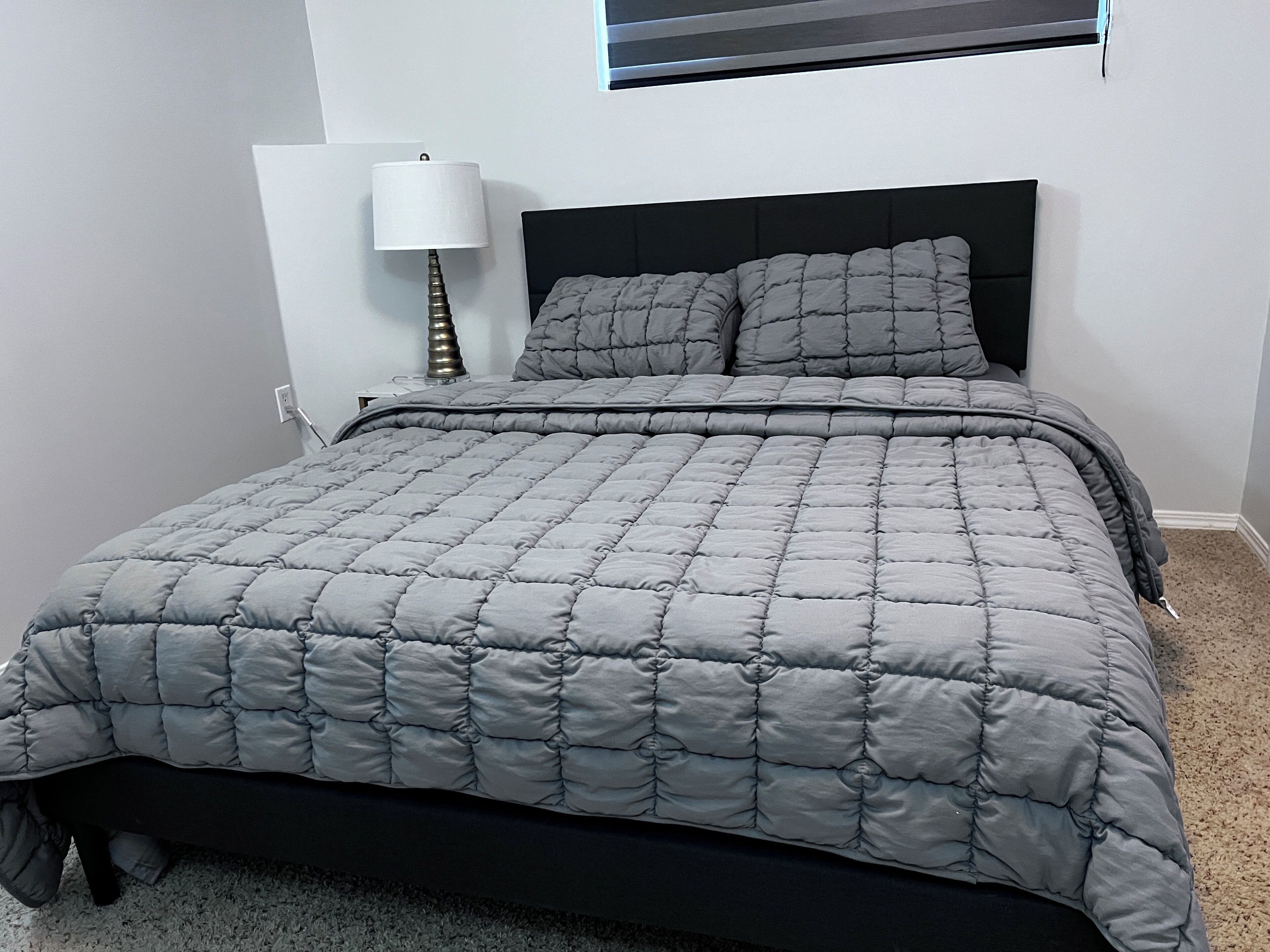 Un lit avec une couvre-lit matelassée grise et une tête de lit noire, dans une pièce aux murs blancs.