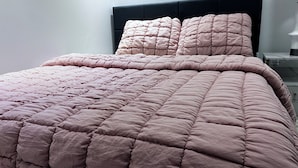 Un lit avec une couette rose et des oreillers assortis, une table de chevet avec une lampe, et un mur blanc.