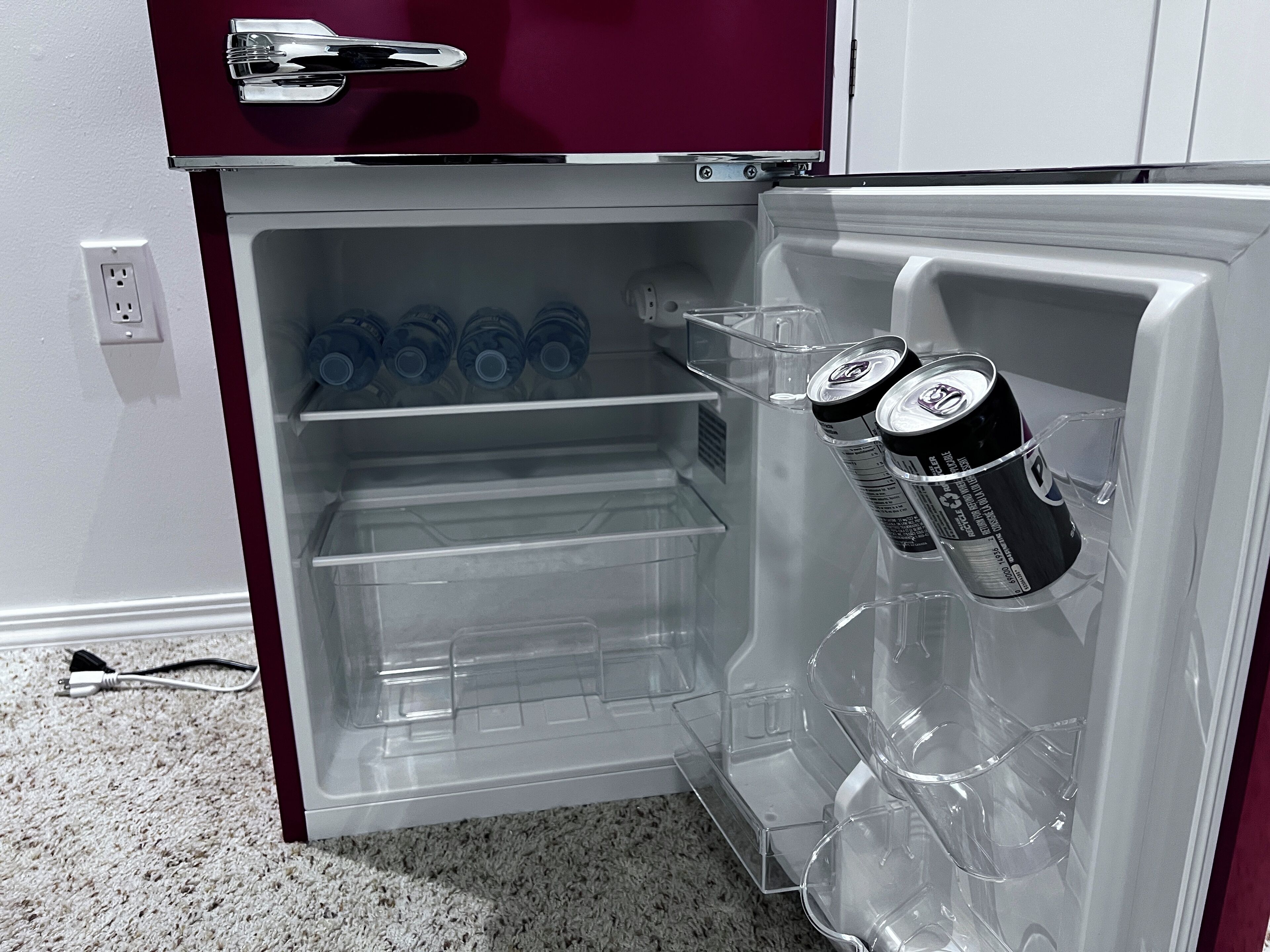 Ein offener Kühlschrank mit klaren Regalen, ein paar Wasserflaschen und zwei Dosen Limonade.