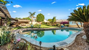 A backyard with a swimming pool, surrounded by various plants and a thatched-roof structure. - Mesa Desert Oasis • Pool • Grill (Mesa)
