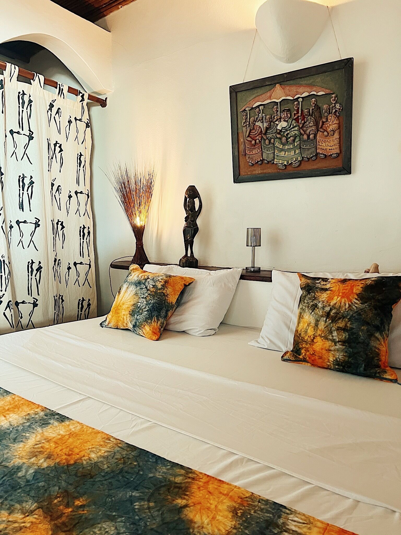 A bedroom with a bed, pillows, a nightstand, a lamp, a wall-mounted painting, and a curtain with a pattern.