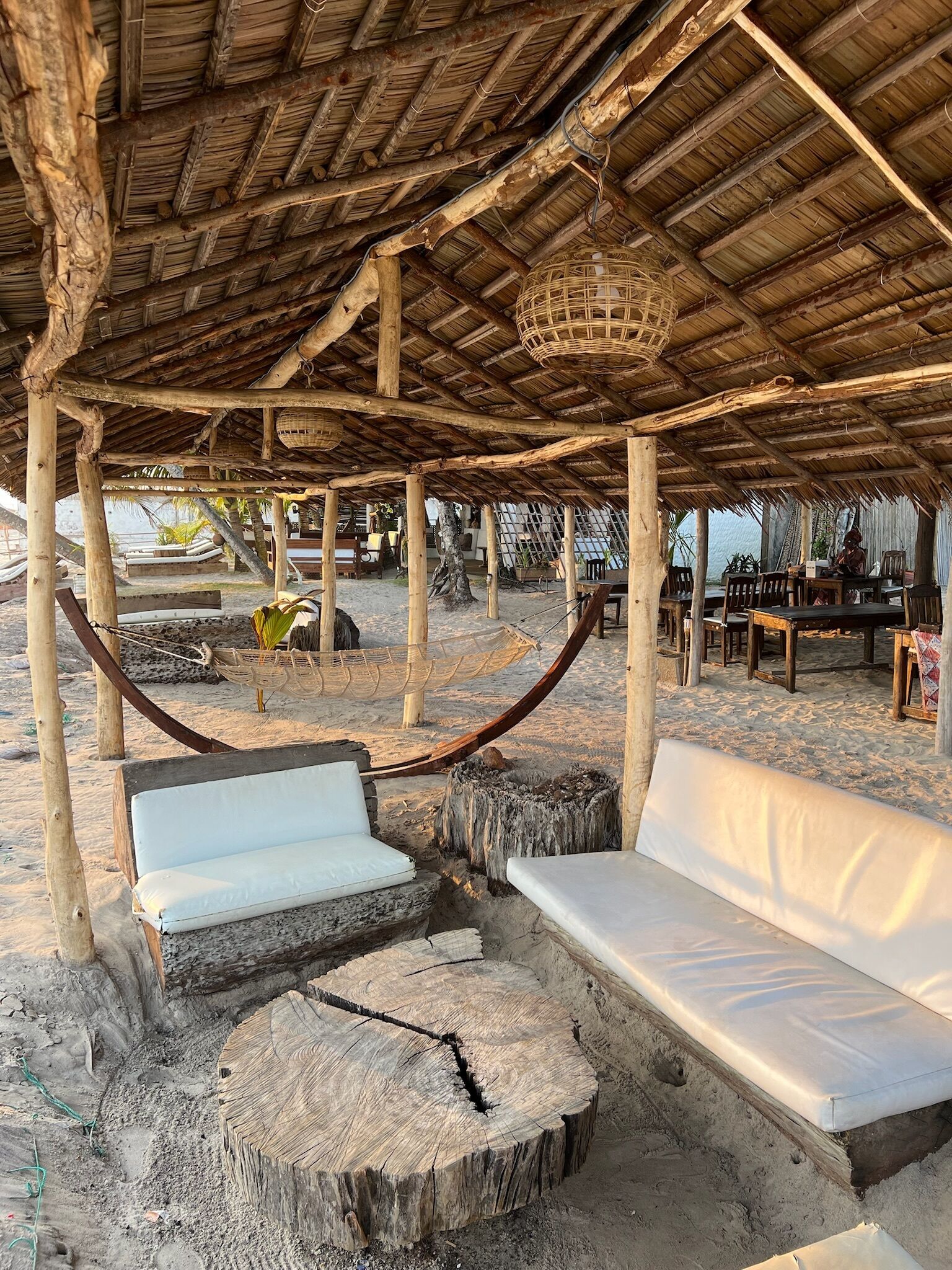 Un área de descanso frente a la playa con mobiliario de madera, una hamaca y una estructura de techo de paja.