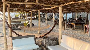 Un área de descanso frente a la playa con mobiliario de madera, una hamaca y una estructura de techo de paja.