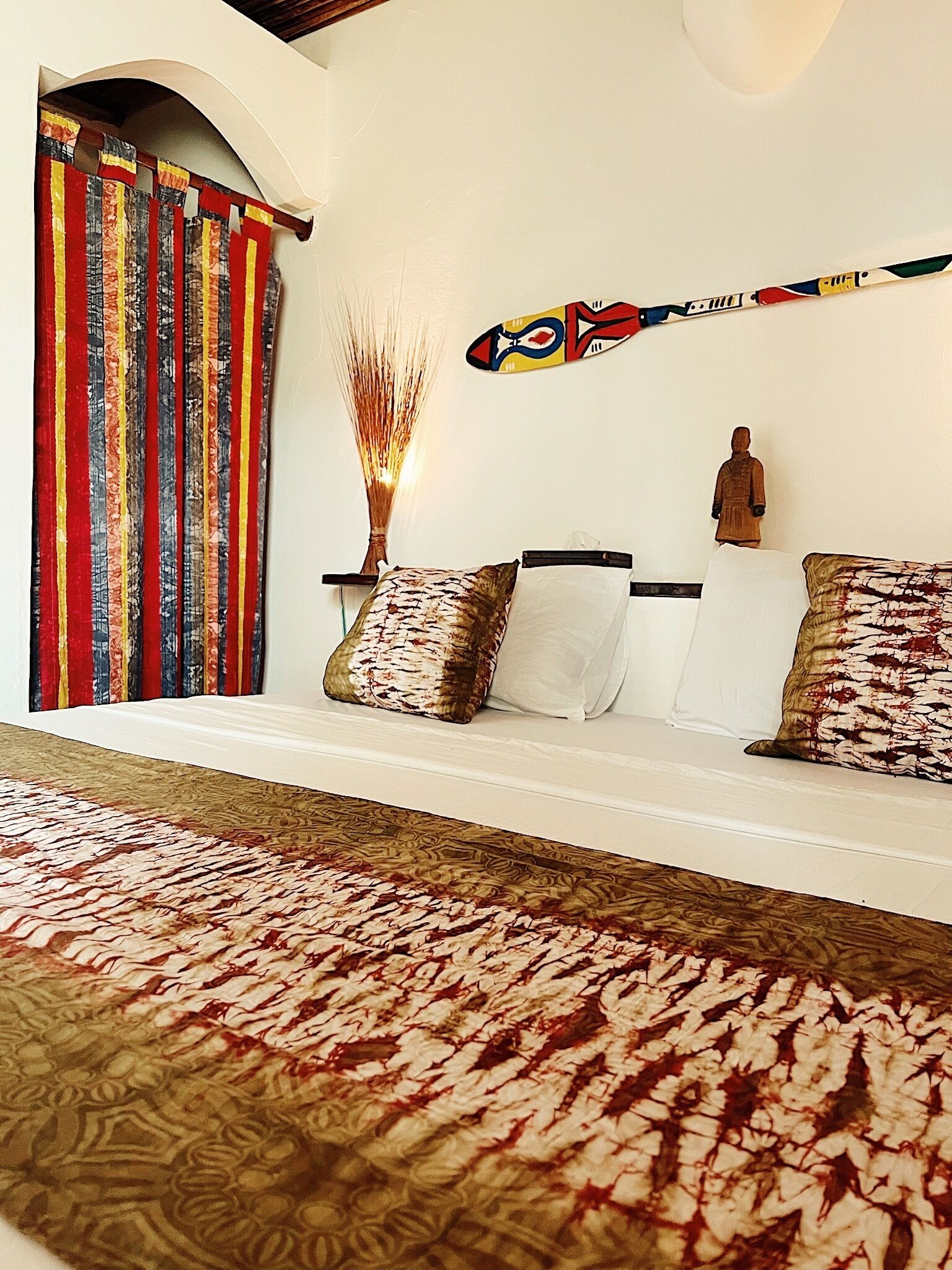 A bedroom with a bed, a bench with cushions, a decorative wall hanging, and a curtain.