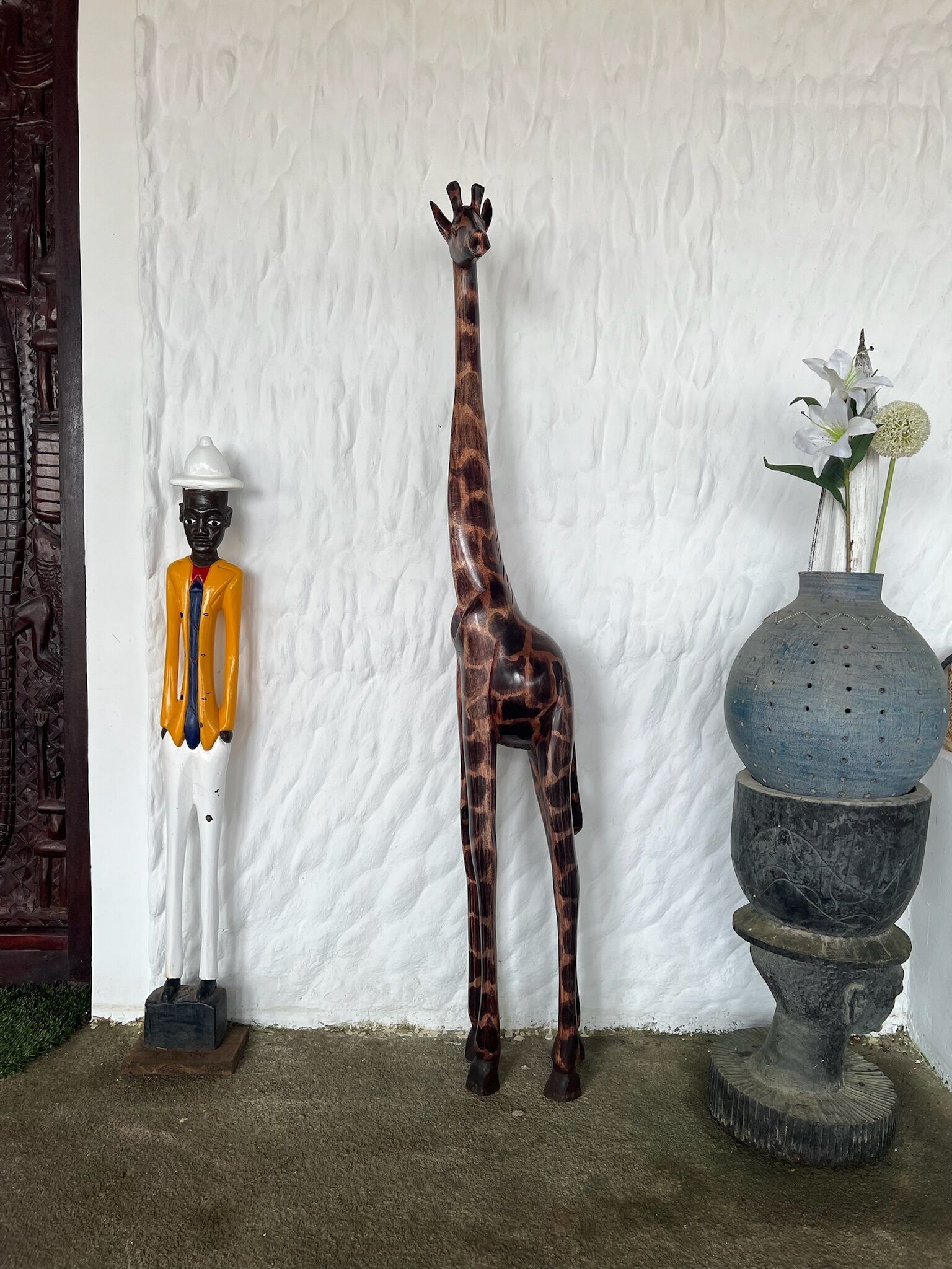 A giraffe sculpture, a human figure, and a vase with flowers against a textured wall.