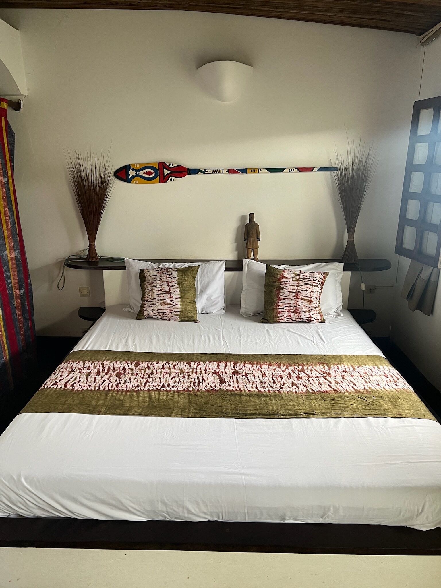 A neatly made bed with white and patterned bedding, a decorative wall hanging, and two tall vases on nightstands.
