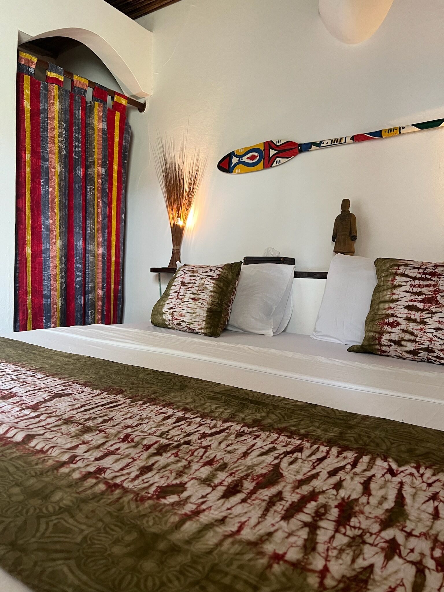 A bedroom with a bed, pillows, a wall-mounted sculpture, and a colorful curtain.