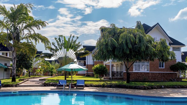 Piscina com cadeiras de descanso e guarda-sóis, cercada por jardins bem cuidados e árvores.