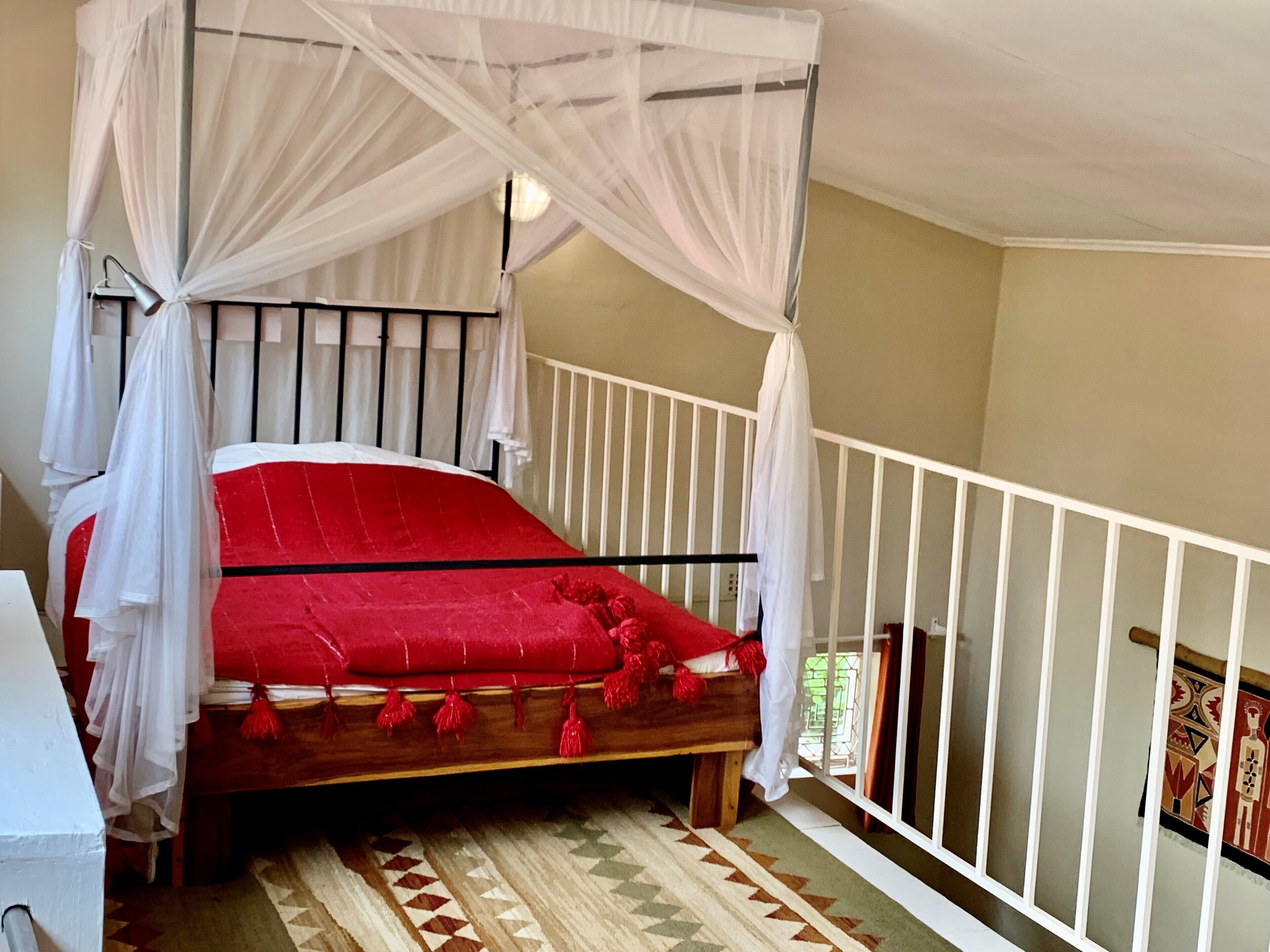 Una habitación con una cama con dosel, cortinas blancas, una colcha roja y una alfombra estampada.