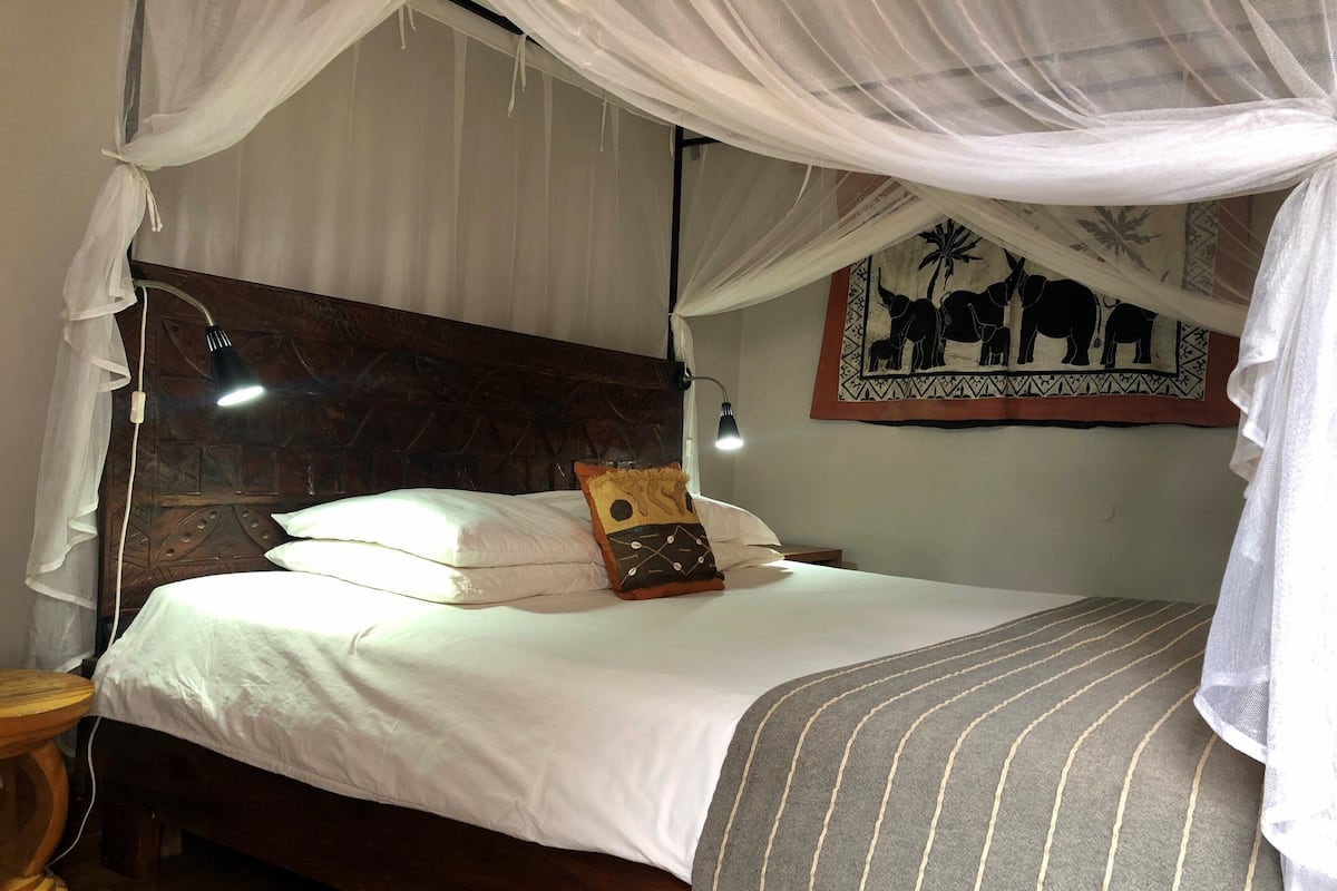 Un lit avec du linge de lit blanc, une tête de lit en bois, un coussin décoratif, une tapisserie murale et une lampe de chevet.