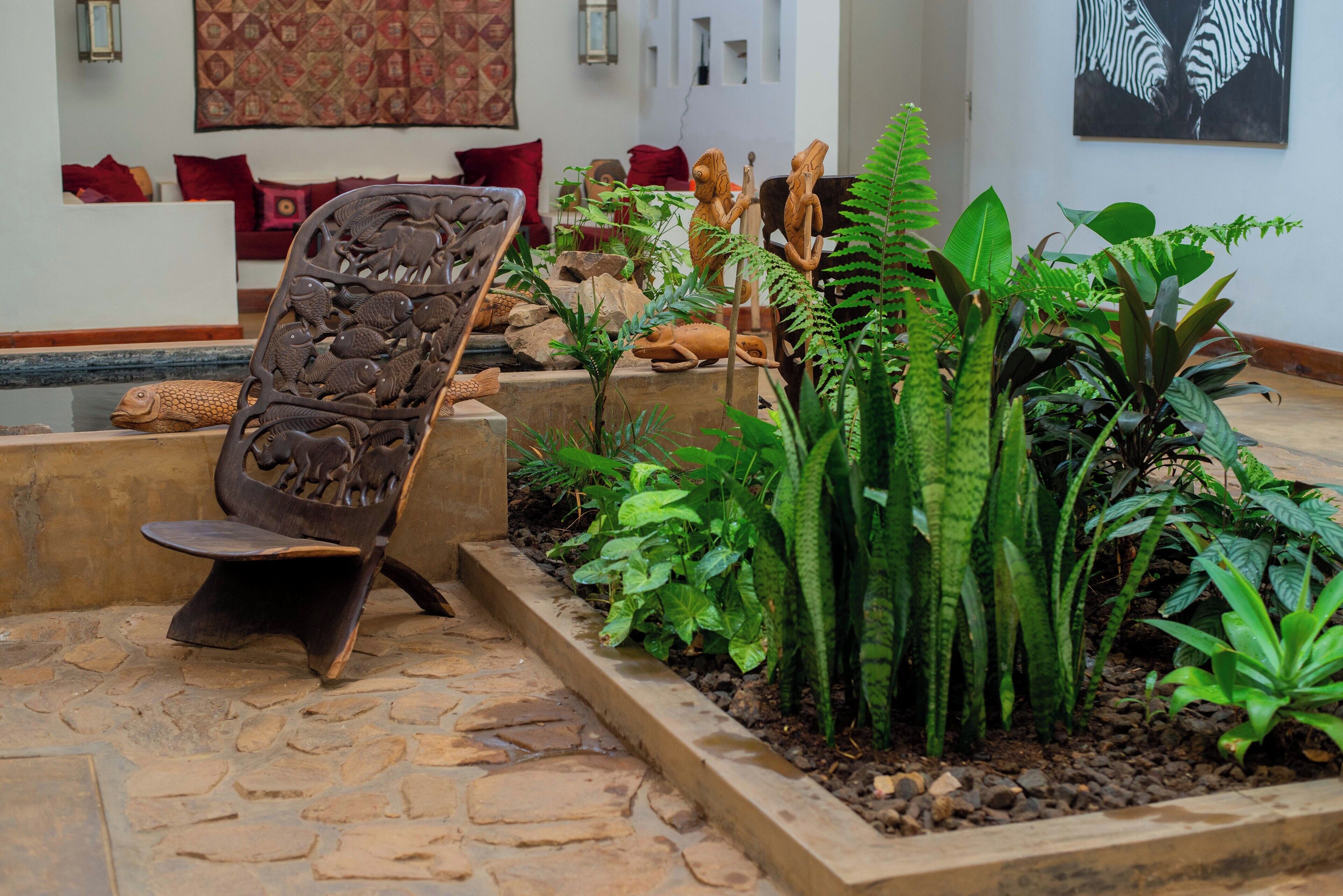 Vestíbulo con suelo de piedra, una silla de madera, un jarrón con plantas diversas y un tapiz rojo con estampado.