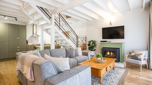 A modern living room with a grey sectional sofa, a wooden coffee table, a fireplace, a TV mounted on the wall, and a staircase leading to an upper floor. - Wavesong on Watts - Pet Friendly with Spa (Callala Beach)