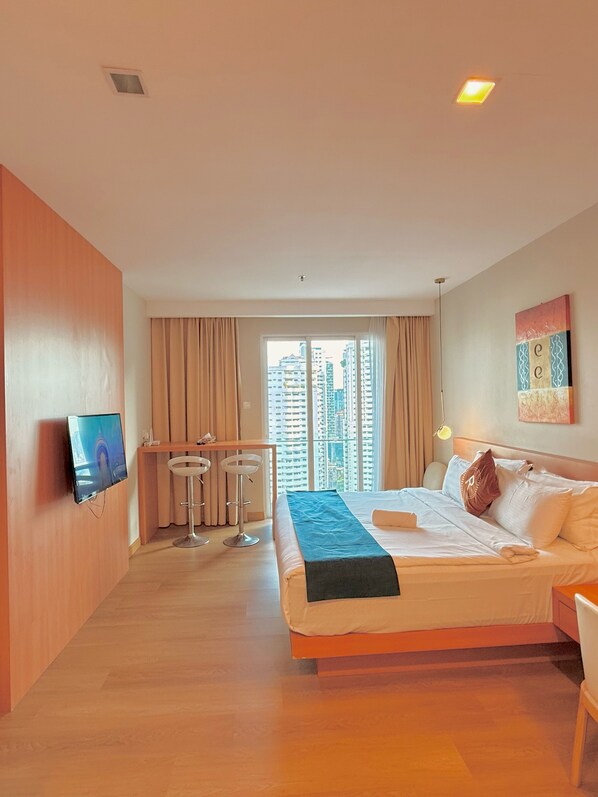 A hotel room with a bed, a desk with two chairs, a TV, and a large window with a city view.