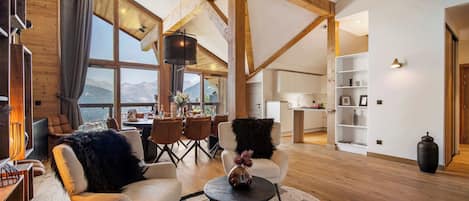 Amplia sala de estar con vigas de madera, un ventanal con vistas a la montaña, zona de comedor y cocina.