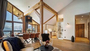 A spacious living room with a high ceiling, wooden beams, a dining area with a mountain view, and a kitchen with white cabinets.