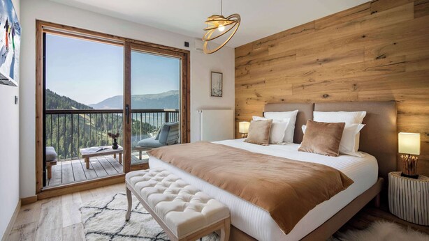 A bedroom with a large bed, wooden headboard, a bench, a nightstand, and a view of a balcony with a chair.