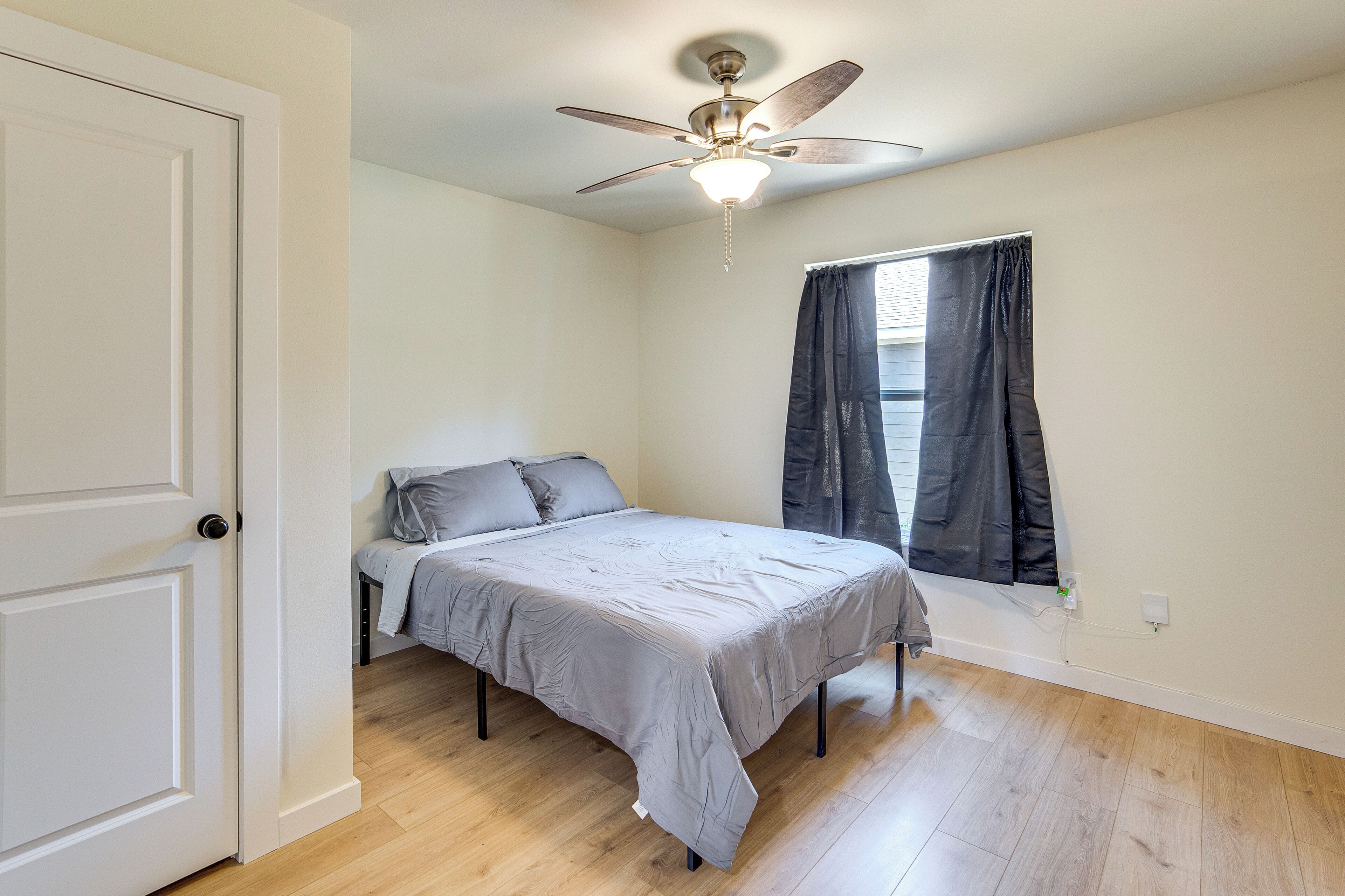A bedroom with a bed, a ceiling fan, a window with curtains, and hardwood flooring.