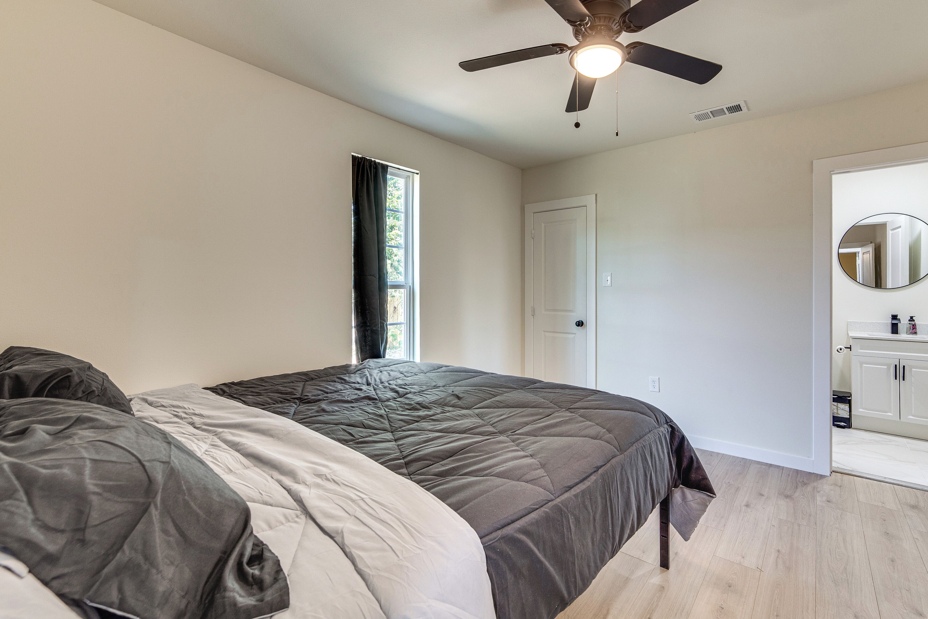 Un dormitorio con cama, ventilador de techo, ventana con cortinas, puerta y baño con espejo y lavamanos.
