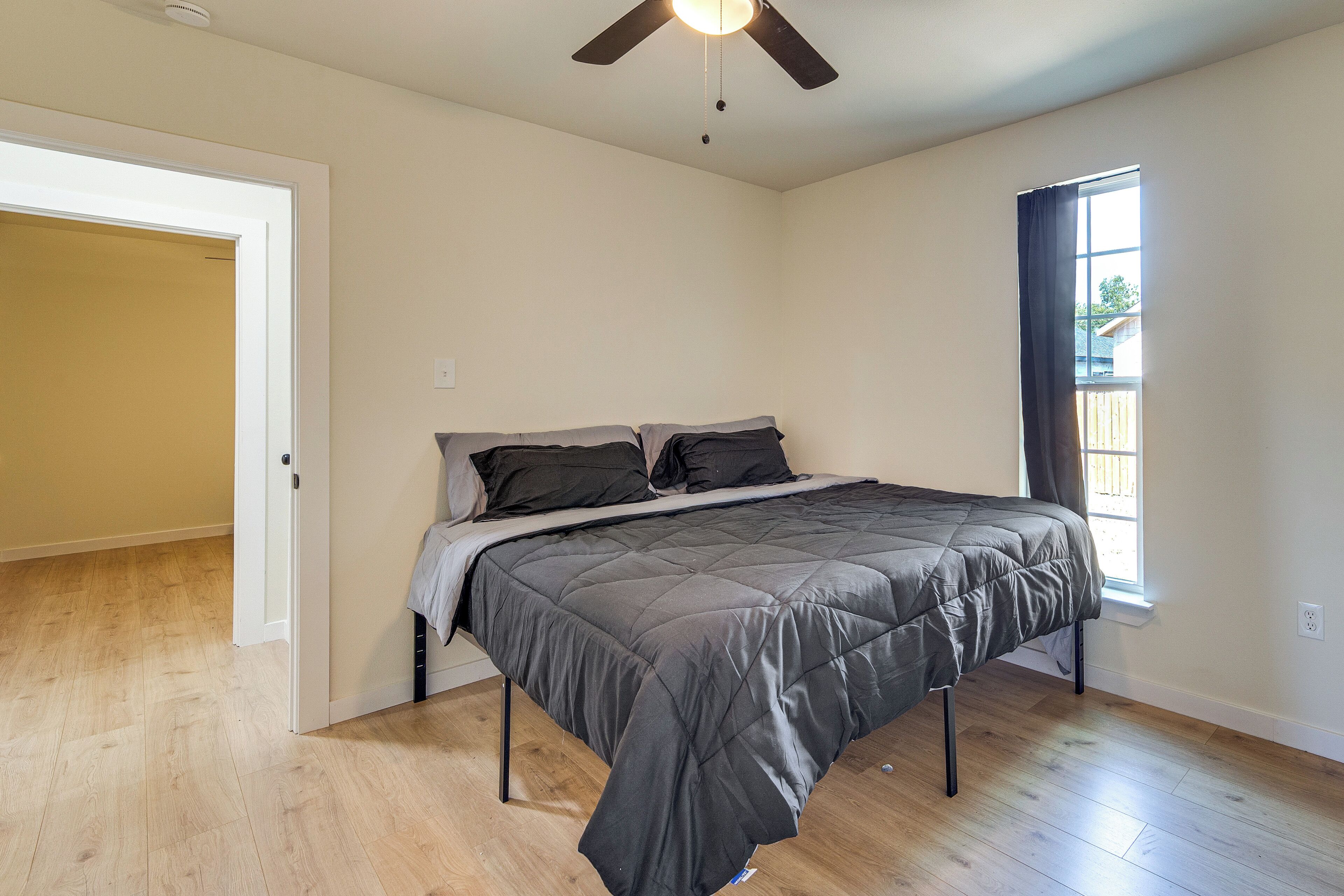 A bedroom with a bed, a ceiling fan, a window with curtains, and a door leading to another room.