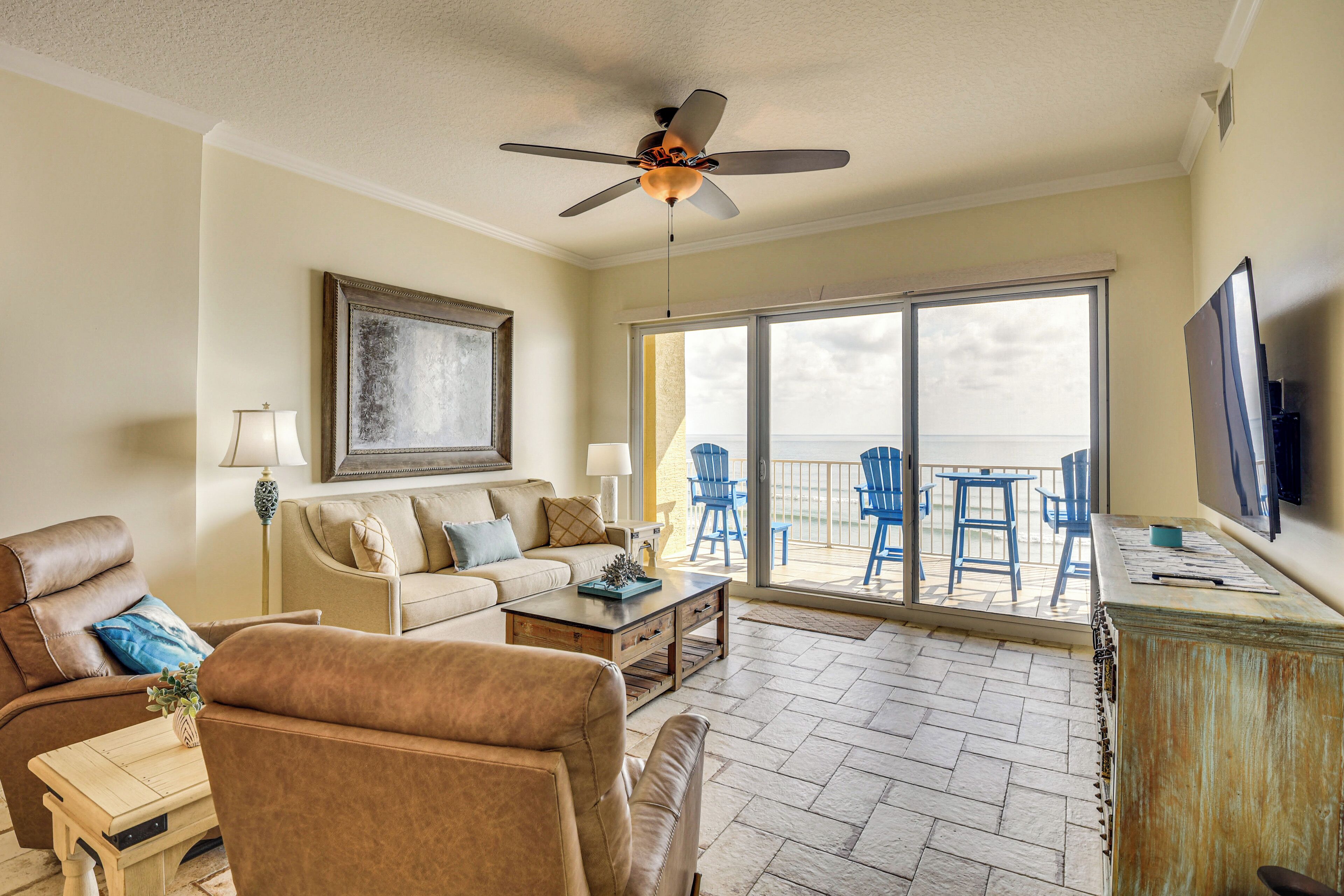 A living room with a sofa, armchairs, a coffee table, and a TV. There is a balcony with chairs and a table.