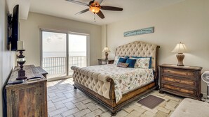 A bedroom with a large bed, a ceiling fan, a nightstand with a lamp, and a view of the ocean through a sliding door. - Direct Beach Access: NSB Condo w/ Community Perks (New Smyrna Beach)