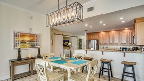 A dining area with a wooden table and chairs, a hanging light fixture, and a kitchen with wooden cabinets and a stainless steel refrigerator. - Direct Beach Access: NSB Condo w/ Community Perks (New Smyrna Beach)