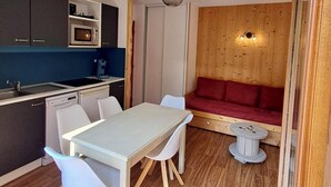 Une cuisine compacte dotée d’appareils électroménagers modernes, un coin repas avec une table et des chaises blanches, et un confortable canapé rouge adossé à un mur en bois.